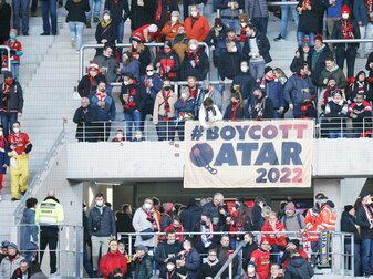 Fans des SC Freiburg stehen auf der Tribüne vor und hinter einem Banner mit der Aufschrift „#BOYCOTT QATAR 2022“. Fans des SC Freiburg stehen auf der Tribüne vor und hinter einem Banner mit der Aufschrift „#BOYCOTT QATAR 2022“.