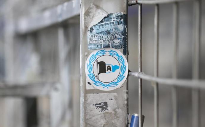 Ein Aufkleber mit dem Logo von Arminia Bielefeld klebt auf einem Zaun. Foto: Friso Gentsch/dpa
