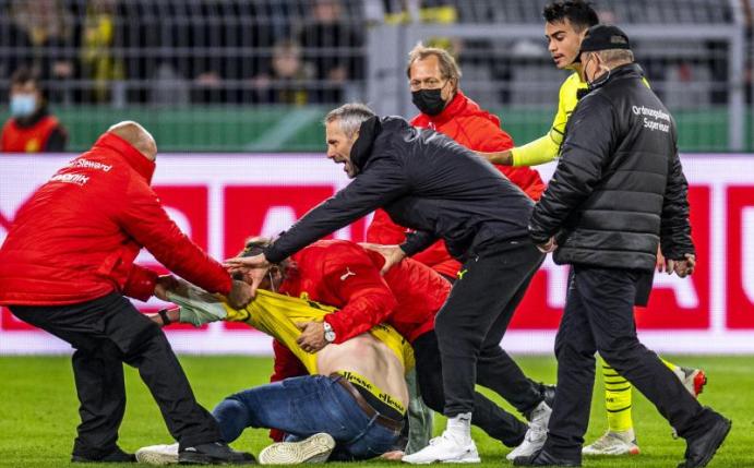 Ordner bringen einen Fan, der auf das Spielfeld gelaufen ist, vom Feld. Foto: David Inderlied/dpa